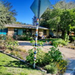 Front view of house and flower beds with a yield sign that a friendly neighbor has woven bright colored stripes of yarn around the pole.