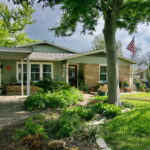 The curb view of 1408 Yorkshire has a curved bed with native grasses and stones. It is surrounding a mature oak tree and has a brick walkway leading to front porch.