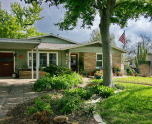 The curb view of 1408 Yorkshire has a curved bed with native grasses and stones. It is surrounding a mature oak tree and has a brick walkway leading to front porch.