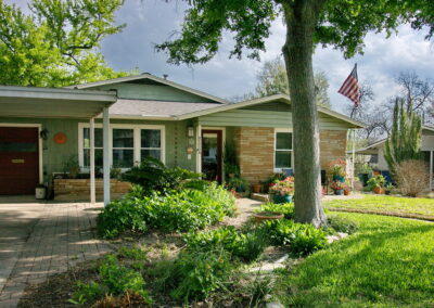 The curb view of 1408 Yorkshire has a curved bed with native grasses and stones. It is surrounding a mature oak tree and has a brick walkway leading to front porch.