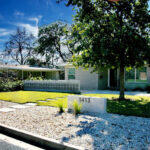 A front curb view of 1413 Ridgemont Dr. This is a lovely example of a minimalist, low water yard. The yard is divided up into a large front gravel section with a concrete wall displaying the numbers to the house. In front of the numbers are two lime green native grasses. Square shaped concrete pavers divide the yard and lead up to the door. The right side of the yard has a large shade tree planted in the grass section. To the left of the house is a beautiful concrete 3 foot dividing wall which contains petal shaped mid-century breeze blocks.