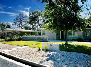 A front curb view of 1413 Ridgemont Dr. This is a lovely example of a minimalist, low water yard. The yard is divided up into a large front gravel section with a concrete wall displaying the numbers to the house. In front of the numbers are two lime green native grasses. Square shaped concrete pavers divide the yard and lead up to the door. The right side of the yard has a large shade tree planted in the grass section. To the left of the house is a beautiful concrete 3 foot dividing wall which contains petal shaped mid-century breeze blocks.