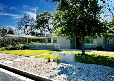 A front curb view of 1413 Ridgemont Dr. This is a lovely example of a minimalist, low water yard. The yard is divided up into a large front gravel section with a concrete wall displaying the numbers to the house. In front of the numbers are two lime green native grasses. Square shaped concrete pavers divide the yard and lead up to the door. The right side of the yard has a large shade tree planted in the grass section. To the left of the house is a beautiful concrete 3 foot dividing wall which contains petal shaped mid-century breeze blocks.