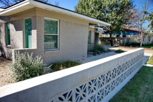 This image displays a close-up view of the Mid-Century breeze blocks and concrete half wall. Behind the wall you can see a bed made of small white limestone pebbles with an evergreen trailing plant and a dwarf silver cloud sage bush.