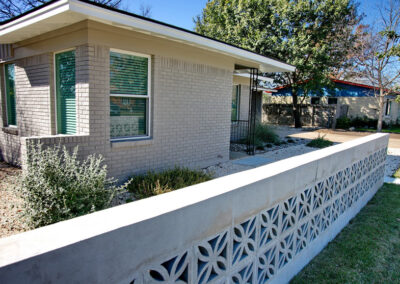 This image displays a close-up view of the Mid-Century breeze blocks and concrete half wall. Behind the wall you can see a bed made of small white limestone pebbles with an evergreen trailing plant and a dwarf silver cloud sage bush.