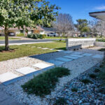 A view taken from the driveway where you'll find the squared paved walkway headed towards the front door which runs perpendicular to the pathway from the middle of yard. The pavers are surrounded by a bed of crushed limestone pebbles. It is adjacent to a river rock bed with runs along side the front of house to the right of the front door. the beds contain trailing rosemary, monkey grass, and blue oat grass.