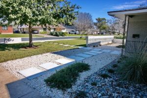 A view taken from the driveway where you'll find the squared paved walkway headed towards the front door which runs perpendicular to the pathway from the middle of yard. The pavers are surrounded by a bed of crushed limestone pebbles. It is adjacent to a river rock bed with runs along side the front of house to the right of the front door. the beds contain trailing rosemary, monkey grass, and blue oat grass.