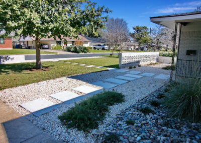 A view taken from the driveway where you'll find the squared paved walkway headed towards the front door which runs perpendicular to the pathway from the middle of yard. The pavers are surrounded by a bed of crushed limestone pebbles. It is adjacent to a river rock bed with runs along side the front of house to the right of the front door. the beds contain trailing rosemary, monkey grass, and blue oat grass.