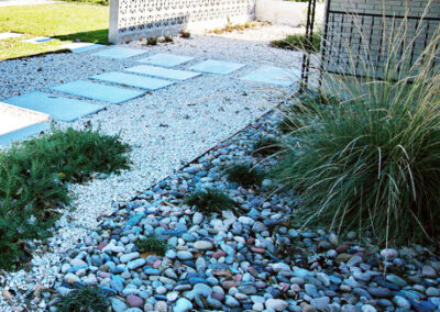 This image depicts a closeup view of the multicolored river rock bed near the front porch. The bed contains several mounded monkey grass, two trailing rosemary plants, and a large Texas Blue stem grass.