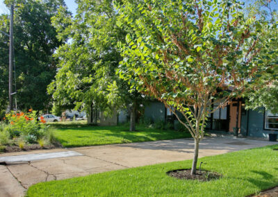 A front view of house from right of the driveway. The image depicts three medium sized shade trees. Also, the front xeriscaped bed is on full display.