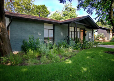 A view of the left side of house with large shaded flower bed. The bed has a white Turk's cap bush, white salvia, white lantana, and black foot daisies. There is also blue and red salvia greggi mixed throughout.