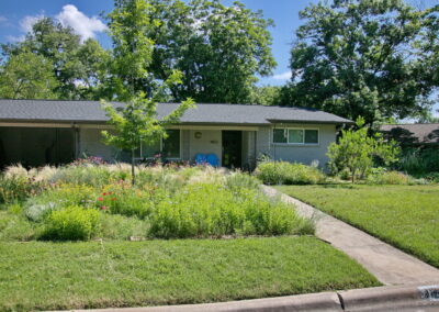 This image depicts a front curb view of 1802 Corona Dr. It has a large meadow bed to the right side of walkway leading to the front door. The meadow is filled with colorful pollinators. You'll find pink coneflowers, orange bulbine, lavender mist flower, yellow lantana, red poppies, and various native grasses.