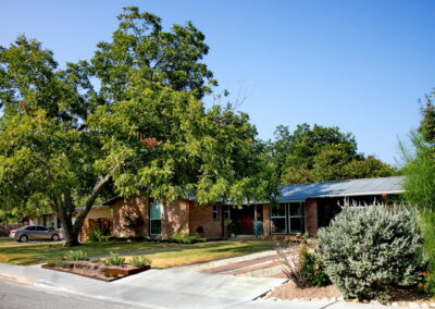 This image depicts the front view of 6008 Belfast Dr. To the left of driveway is a large Pecan Tree which provides shade over much of the yard. The front sidewalk is anchored by a two tier steel planter box to the right of concrete walkway. It is filled with gopher plants, silver pony foot, and various hardy succulents.