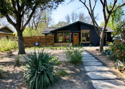 A front view of 1419 Westmoor Dr. with a limestone walkway leading to the Navy painted home with a bright orange door. Along the walkway you'll find decomposed granite beds with yuccas and agaves.