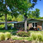 A street view of the house depicting two oak trees surrounded by rock lined beds with native grasses and red salvia greggi.