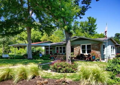 A street view of the house depicting two oak trees surrounded by rock lined beds with native grasses and red salvia greggi.