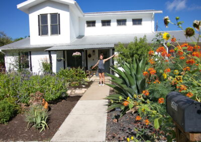 This image shows the owner of 5400 Wellington standing proudly amongst her beautiful front yard garden. You'll find a large blue agave, several blue salvias, canna lilies infant of the porch, and more colorful Pride of Barbados.