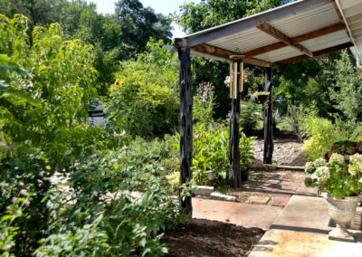 This image depicts the overhang structure added on to the front porch. It is has a metal cover and natural wood columns. In front of the porch is a cluster of small red cannas. hanging from the structure is a flower pot full of purslane and a small wind chime.