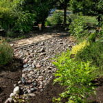 A side bed filled with various colors of river rock. There is also a yellow bell, orange salvia greggi, coral yucca, and a sweet william.