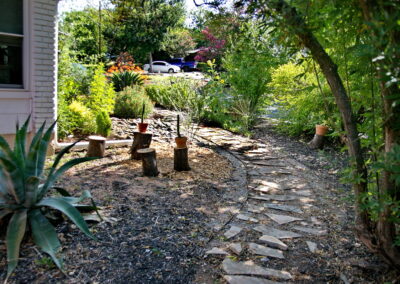 A view of a stone walkway winding around the house. Along the trail is a large blue Agave. There are cut off tree stumps with small flower pots on top of each one.