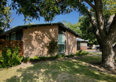 This image shows the shade area against the left side of home. You'll find a steel container box across the width of the peach brick house. It contains Fastia Japonica, lirope grasses, a small Japanese maple, and river fern. There is also a large Turks cap with small red flowers planted to the right side of container box.