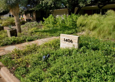 A view of a gray brick signage with the house address and it is surrounded by trailing yellow lantana.