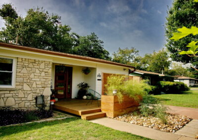 Another view of the front porch which has been extended as a deck. The deck has two lounge chairs on each side of the front door. Potted plants are also displayed.