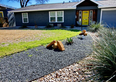 A view of the curved flower bed with dark gray gravel leading to the front door. The gravel has large rocks placed along the curve and includes a blue agave and coral yuccas.
