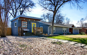 This image depicts the guest house which is to the left of the main house on the property. In front of the house is a large yard with multi colored river rock and concrete square steps which lead to the bright yellow front door.