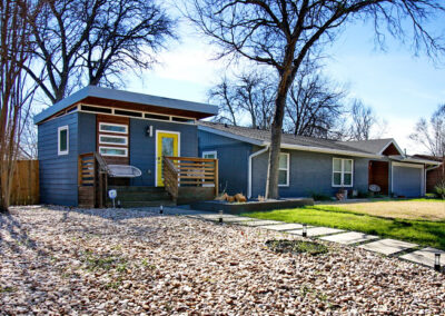 This image depicts the guest house which is to the left of the main house on the property. In front of the house is a large yard with multi colored river rock and concrete square steps which lead to the bright yellow front door.