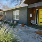 A close up view of the front yellow door with a white woven chair on the front porch. You'll also find white rock flower beds along the exterior of the house with native grasses and planter boxes containing cacti.