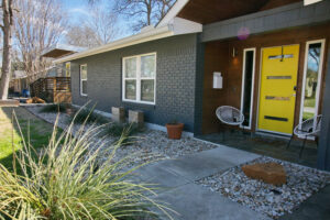 A close up view of the front yellow door with a white woven chair on the front porch. You'll also find white rock flower beds along the exterior of the house with native grasses and planter boxes containing cacti.