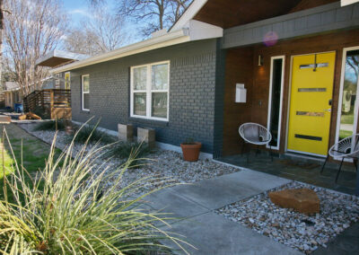 A close up view of the front yellow door with a white woven chair on the front porch. You'll also find white rock flower beds along the exterior of the house with native grasses and planter boxes containing cacti.