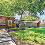 Front curb view of 5207 Robinsdale. It is a gray house with a bright yellow door and a side guest house also with a bright yellow door. The landscaping consists of several xeriscaped beds with gray gravel low water grasses and yuccas. There is also large spaces of green grass.