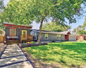 Front curb view of 5207 Robinsdale. It is a gray house with a bright yellow door and a side guest house also with a bright yellow door. The landscaping consists of several xeriscaped beds with gray gravel low water grasses and yuccas. There is also large spaces of green grass.