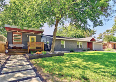 Front curb view of 5207 Robinsdale. It is a gray house with a bright yellow door and a side guest house also with a bright yellow door. The landscaping consists of several xeriscaped beds with gray gravel low water grasses and yuccas. There is also large spaces of green grass.